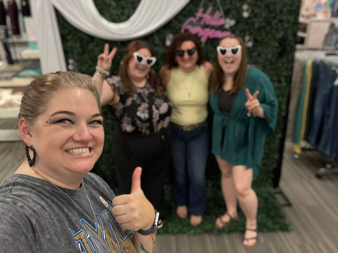 4 women giving thumbs up or peace sign in front of a foliage textured backdrop for selfies